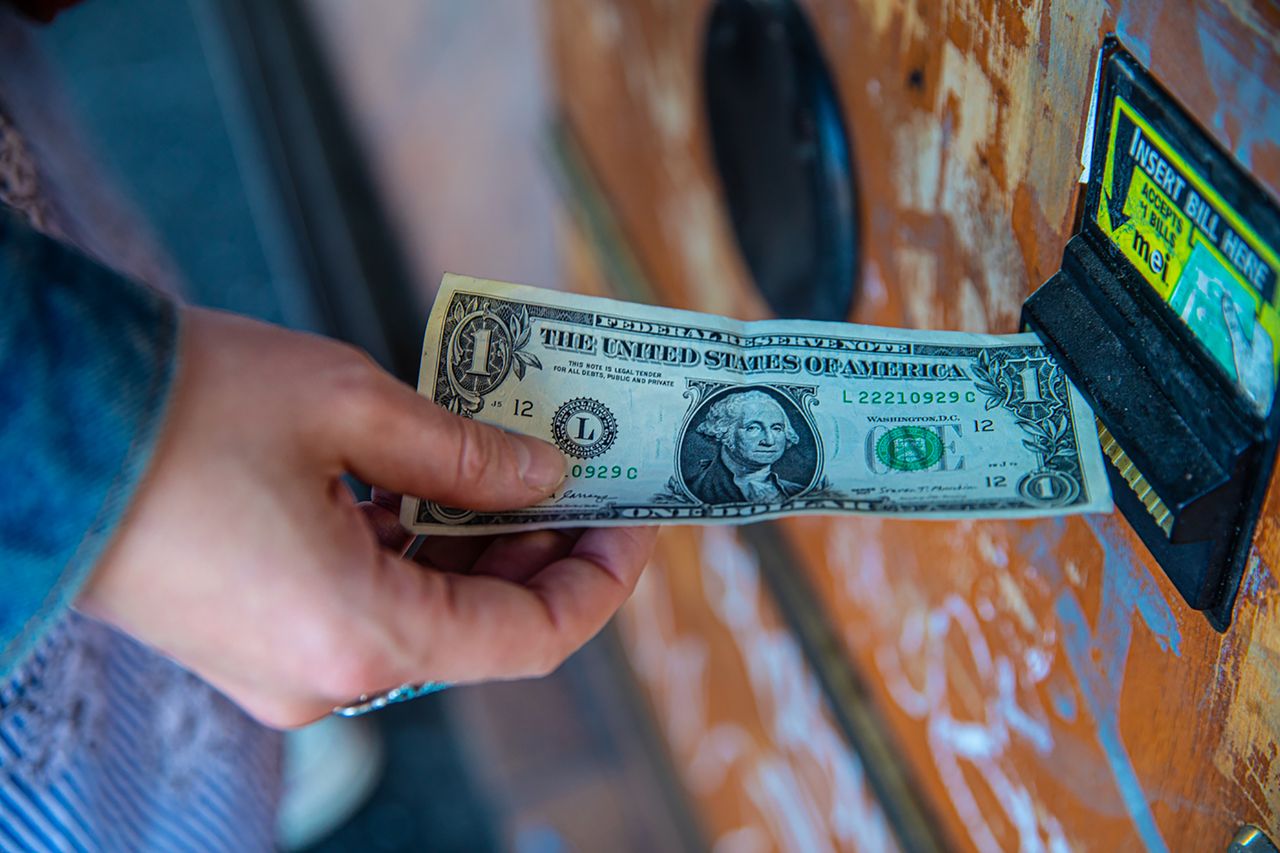 A person inserting a US dollar bill into a vending machine slot, capturing a financial transaction moment.